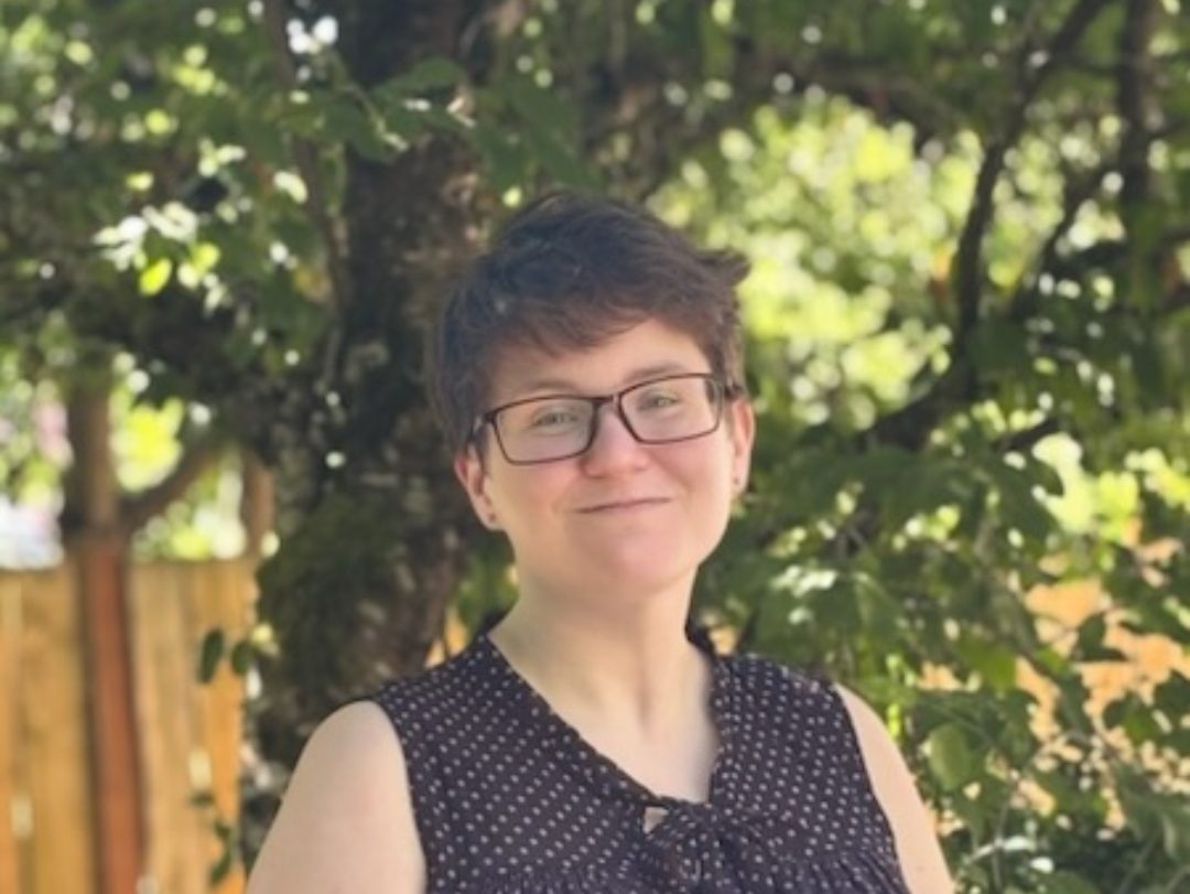 Photo of a white woman with short brown hair in a blue shirt standing in front of trees and fences.